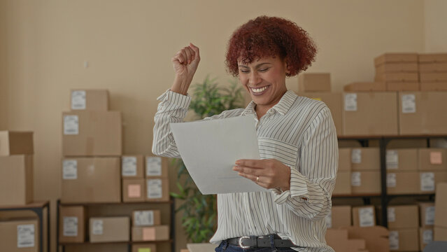 Woman holding document, hand raised in celebration while smiling and reading among stacked cardboard boxes in a building; small business success joy.