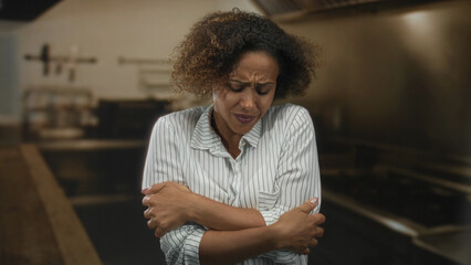 Fototapeta premium Woman hugging her arms across chest in a kitchen building, eyes closed and wincing; discomfort tension.