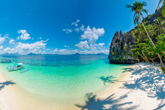 Entalula Beach in El Nido, Philippines, limestone cliffs, vibrant tropical vegetation, and traditional outrigger boats floating on crystal-clear turquoise waters under sunny blue sky of Palawan island