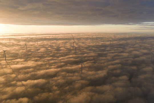Aerial view of Windpark Fryslan wind turbines emerging from a dense layer of golden clouds during sunrise in Breezanddijk, Friesland, Netherlands.