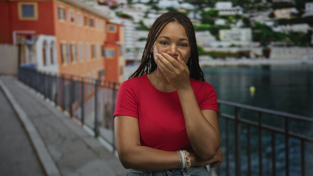 Woman with hand covering mouth on a seaside street promenade near railing and water, braided hair and red tshirt; surprise wonder.