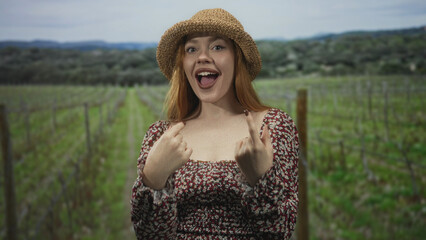 Obraz premium Young redhead woman wearing a straw hat and floral dress shows both middle fingers with tongue out in a vineyard; playful defiance.