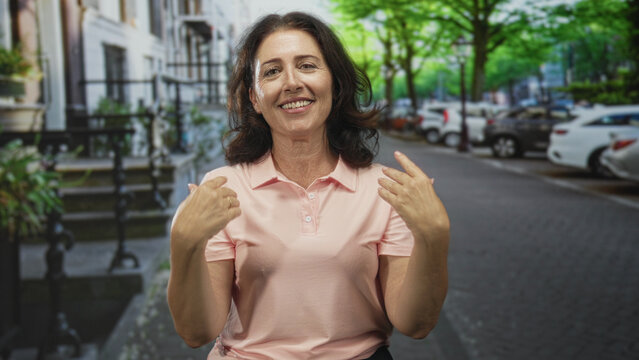 Woman in pink polo pointing both hands toward camera on a tree lined street with parked cars and stoop steps; invitation friendly.