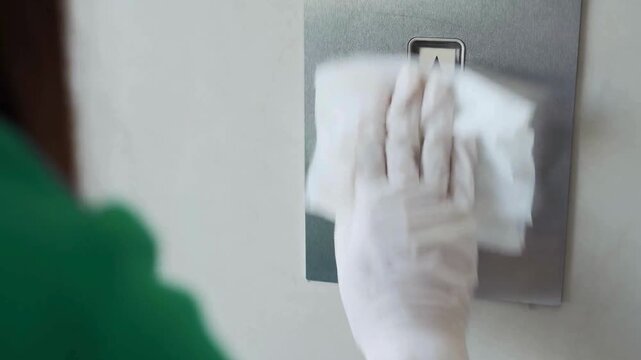 Staff member wearing protective gloves disinfecting elevator buttons using spray bottle and wipe to prevent virus spread in public office building area.