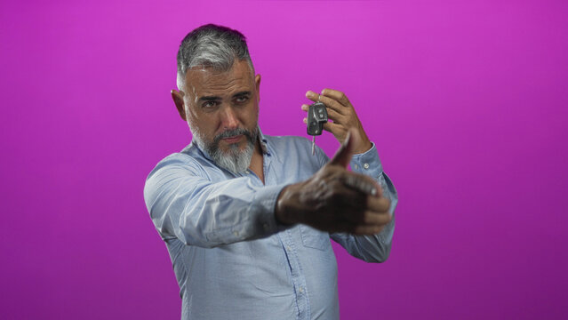Middle aged man holding car key fob between fingers and pointing with thumb in a magenta studio; doubt decision.
