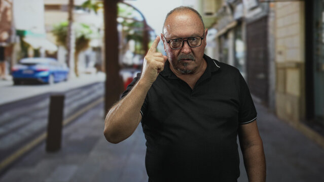 Senior hispanic man wearing glasses and a black polo points finger to temple while standing on a narrow street with bollard, blurred car and storefront; warning.