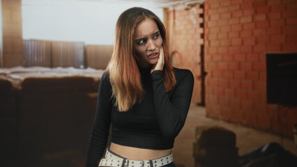 Fototapeta premium Woman hand to cheek with grimace, touching jaw and glancing sideways in unfinished brick building with mattress and window light; anxiety unease isolation.