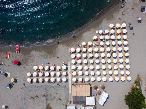 Aerial view of organized rows of white beach umbrellas and sunbeds on the sandy shore with people swimming in the clear blue sea, San Lorenzo, Liguria, Italy.