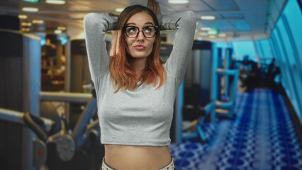 Fototapeta premium Woman with arms raised, hands behind head and exposed midriff in airport terminal, red hair and glasses wearing grey crop top and casual pose; playful ease.