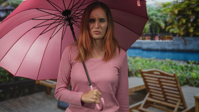 Woman holding pink umbrella, gripping handle and frowning by pool chairs at building pool deck; annoyance solitude.
