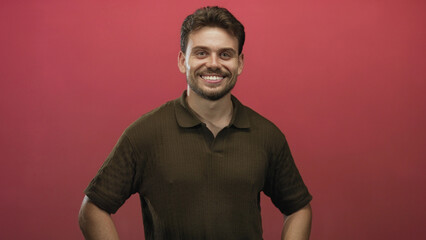 Obraz premium Man smiling with hands on hips in red studio, wearing olive polo and facing camera with relaxed posture; confidence.