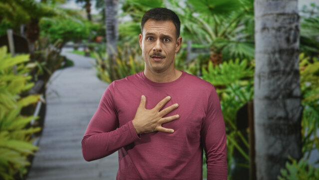 Hispanic man with mustache, hand on chest on boardwalk path, wearing maroon long sleeve shirt and looking earnest; sincerity gratitude.