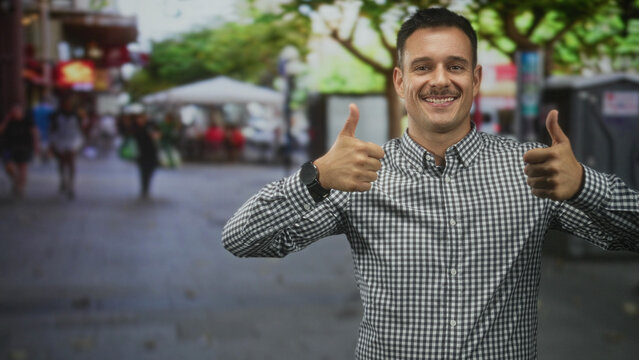 Hispanic man with moustache wearing a checkered shirt, smiling and giving two thumbs up on a busy street lined with outdoor cafes and trees; cheerful.