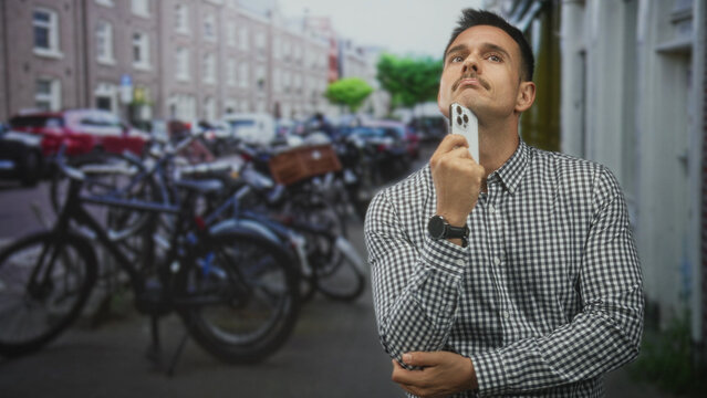 Young hispanic man holding smartphone near chin while looking up on a city street; thoughtful planning.