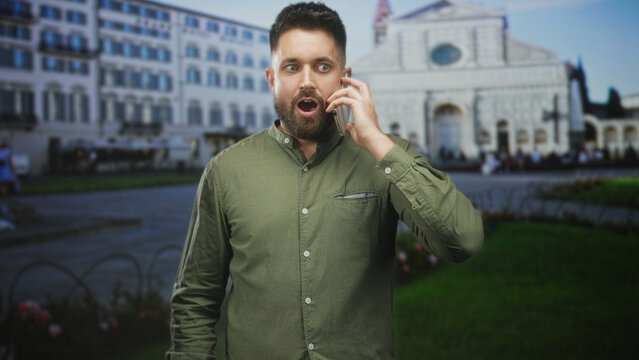 Man holding smartphone to ear, mouth open in surprised gasp while talking before a historic building plaza and cathedral facade; surprise curiosity.