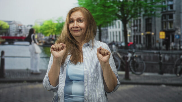 Woman holding both hands in a nervous airquote gesture while standing on a cobbled city street beside a canal bridge; uncertainty apology.