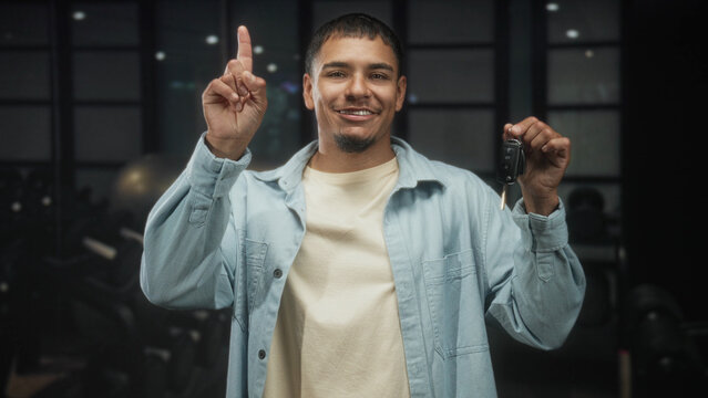 Man holding car keys and pointing finger in studio, smiling and showing the key fob while wearing a denim jacket and t shirt; confidence celebration new car.