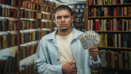 Man holding a fan of us dollars, hand on stomach for stomachache, gaze worried amid bookshelves in a library building  financial concern. © Krakenimages.com