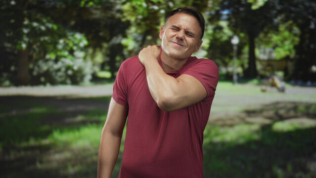 Handsome hispanic man in red polo rubbing his left shoulder and wincing on a sunlit forest path; discomfort recovery.