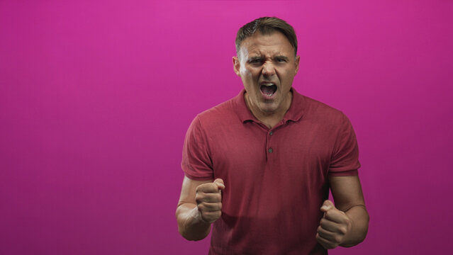 Man cupping his hand to his mouth and shouting against a magenta studio backdrop while wearing a maroon polo, expressing intense vocal outburst; frustration.