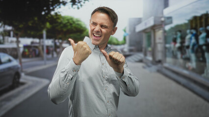 Fototapeta premium Man pointing both thumbs at his chest while winking and grinning, buttondown shirt visible, standing on a city street by a storefront with mannequins and a parked car; confidence.