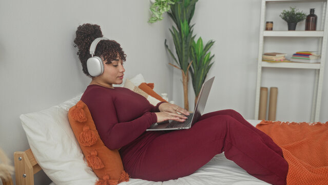 Woman points finger to laptop while wearing headphones and smiling on bed in studio with orange throw and potted plant; relaxation focus.
