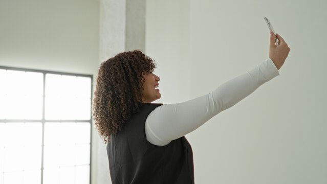 Woman smiling holding smartphone with hand raised for selfie in empty building room with tall window wearing black vest and white top; self confidence.