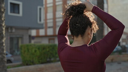 Woman tying hair with hands on street near building and hedge, wearing maroon long sleeve shirt and hoop earrings  joy. © Krakenimages.com