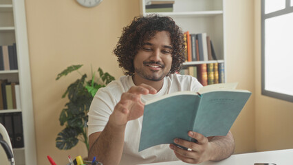 Man reading a paperback book, smiling and turning a page at a desk in a library building with bookshelves and a plant  serenity. © Krakenimages.com
