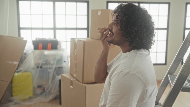 Man holding smartphone to mouth speaking and smiling amid stacked cardboard moving boxes and ladder inside building; moving day joy.