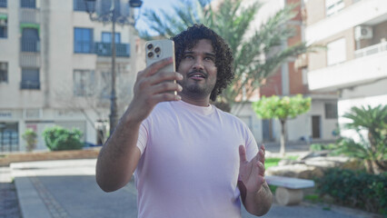 Fototapeta premium Young indian man with curly hair holds smartphone, makes ok sign and smiles on street by building, wearing pink t shirt and gesturing with hand; connection joy spontaneity.