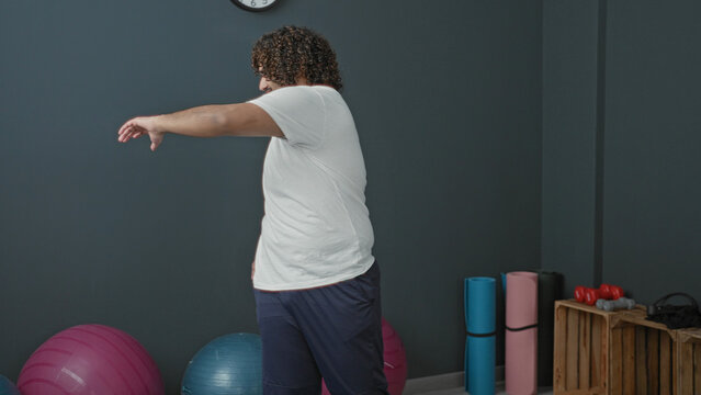 Man stretching side with hand on hip and extended arm in studio gym, surrounded by mats, exercise balls and dumbbells; serenity focus.