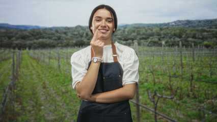Obraz premium Woman in apron touches chin with hand, arms folded, standing among vineyard rows in forest setting with distant hills and olive trees; calm thinking.