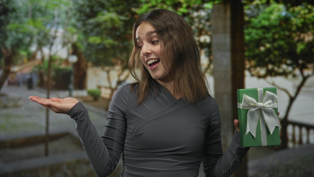 Woman presenting green wrapped gift with palm up gesture on a tree lined street beside stone railing; generosity surprise.