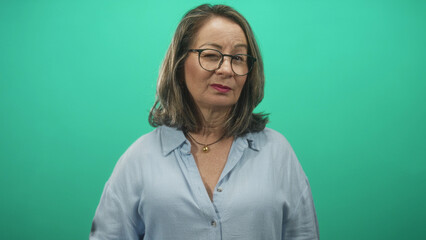 Fototapeta premium Woman pressing finger to lips in a shushing gesture wearing glasses and blouse against a green studio backdrop; silence.