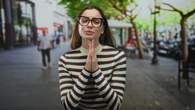 Woman with hands pressed together in pleading pose on a city street lined with trees and cafes; apologetic plea.