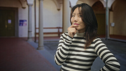 Obraz premium Woman clapping hands with tongue out and closed eyes in building courtyard, wearing striped sweater; playful.