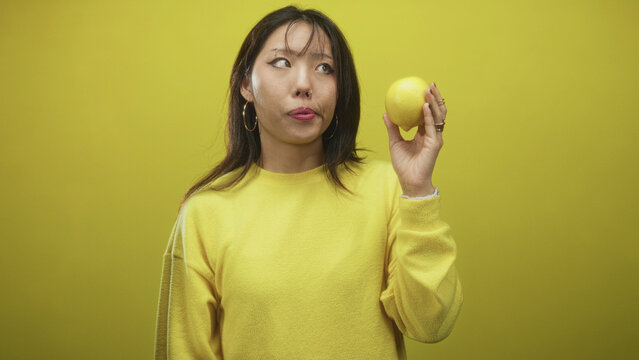 Woman holds lemon near face with raised hand, puckered lips and sideways glance, wearing yellow sweater and hoop earring against seamless yellow studio backdrop; skepticism.