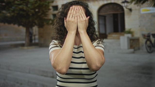 Woman covers face with hands on street outside building entrance before pulling them away to smile broadly; joy.