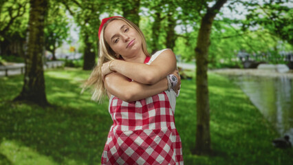 Fototapeta premium Woman chef wearing red check apron and headband hugs her shoulders while smiling by a pond in a forest; homey contentment.