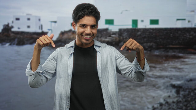 Man pointing both thumbs at his chest in front of a coastal white building with rocky shore and sea; confidence casual.