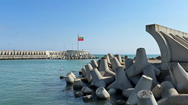 Clear sky over the water at a harbor showing a flag and concrete blocks on the shore. Waves gently touch the structures while a few birds are seen nearby.