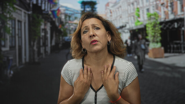 Woman points finger to palm showing empty hand asking for money on a city street lined with shops and rainbow flags; pleading need.