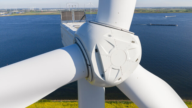 Aerial view of a white wind turbine nacelle and rotor hub overlooking a blue waterway with a passing barge and distant turbines in Ooltgensplaat, Zuid-Holland, Netherlands.