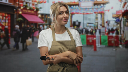 Obraz premium Woman gardener holding trowel with arms crossed on crowded street market in chinatown; local pride contentment.