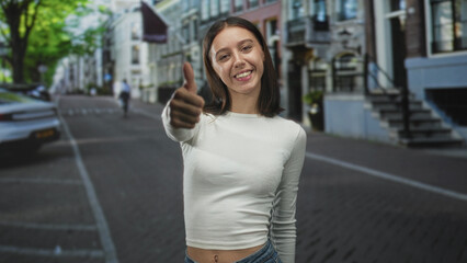 Fototapeta premium Young hispanic woman smiling and giving thumbs up with bare thumb visible on a street lined with buildings; confidence joy.