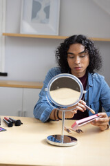 Non-binary adult applying makeup at home at light wood table using mirror, pink palette and brushes © wavebreak3