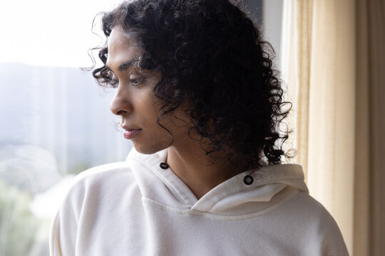 Non-binary adult standing by window wearing cream hoodie with metal grommets, gazing in soft light