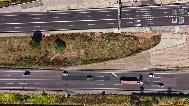 Vehicles move along the multiple lanes of a highway while a patch of grass lies in the middle. The sun shines on the road and surrounding landscape.