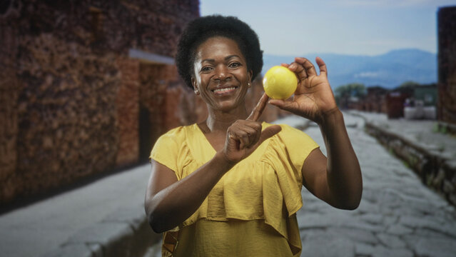 Woman smiling while holding a lemon with raised hand and pointing finger on a sunlit cobblestone street with stone buildings visible; joy health.